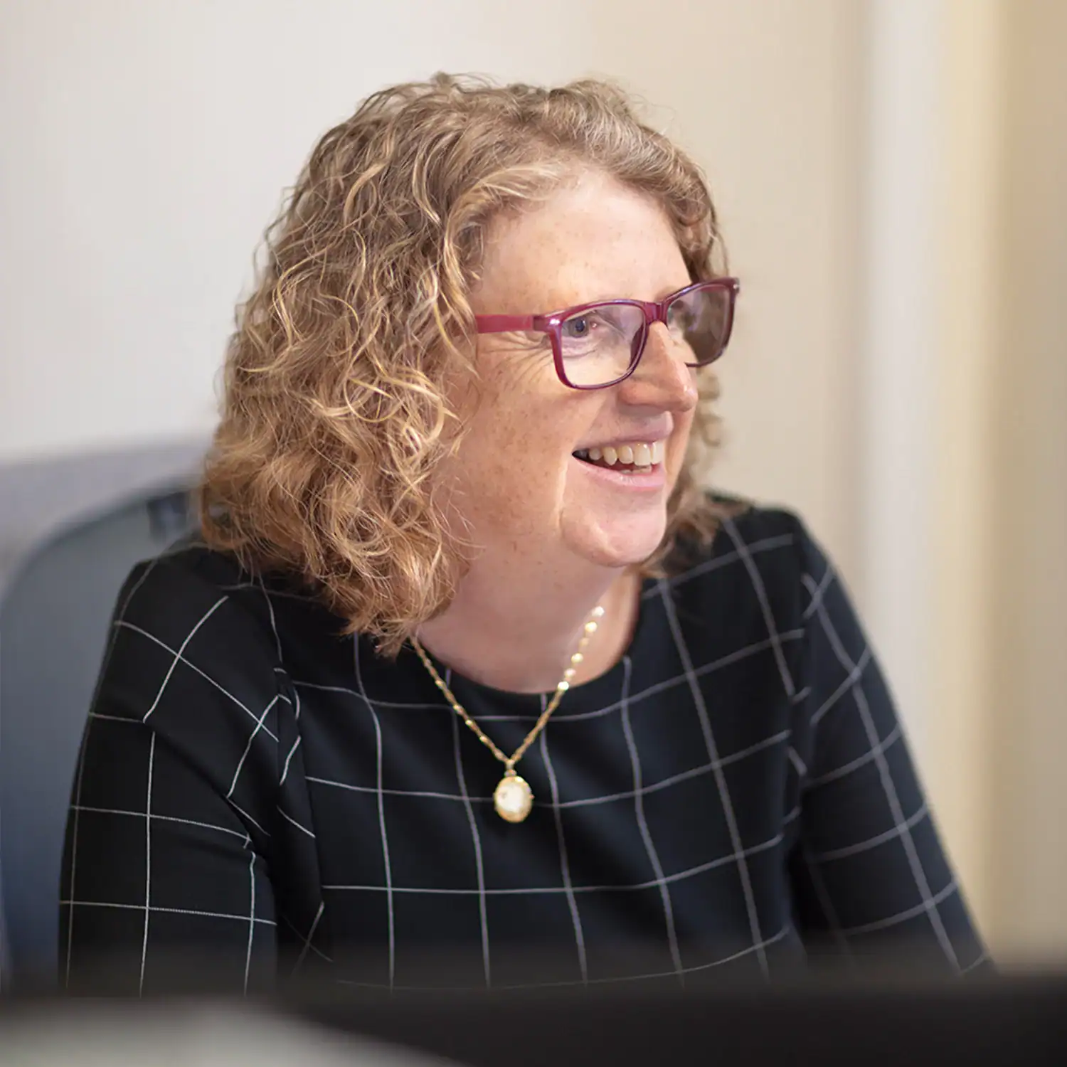 Smiling Woman with Curly Blonde Hair and Pink Glasses, Wearing a Black Grid-pattern Dress and a Gold Pendant in an Office Setting